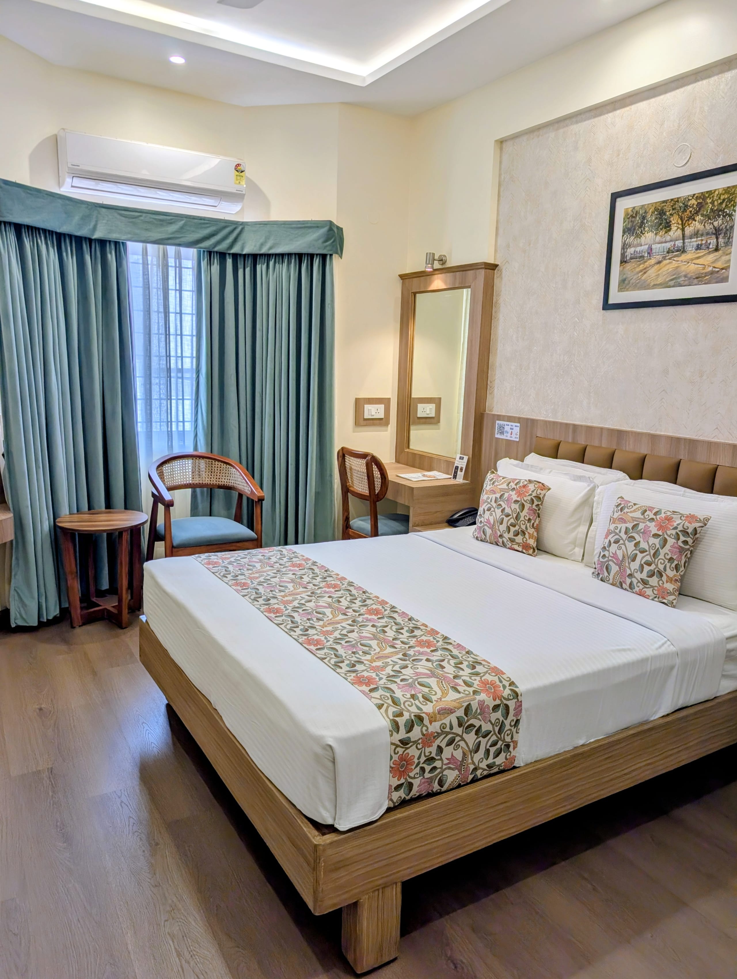 A Gold Superior Room at Bangalore Gate Hotel & Conferences, featuring a double bed with a red bed runner, gold leaf wall art on a grey headboard panel, a ceiling fan, a wall-mounted flat-screen television, teal curtains, and a wooden writing desk.