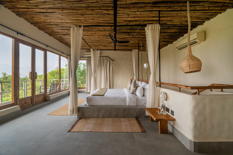 A bedroom with a curtained bed and a full wall of windows onto the forest