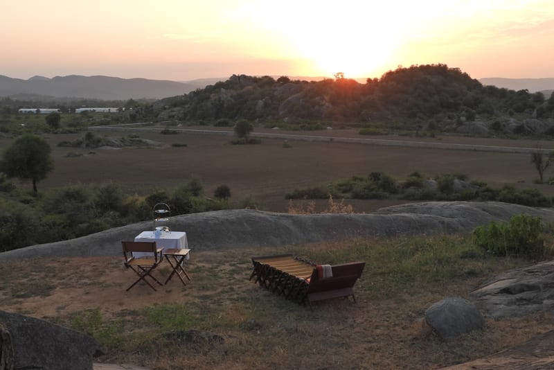 An image of camping site where a chair and table is placed near a lounger bed, and the sun is also setting at the horizon - Utsav Camp Sariska.
