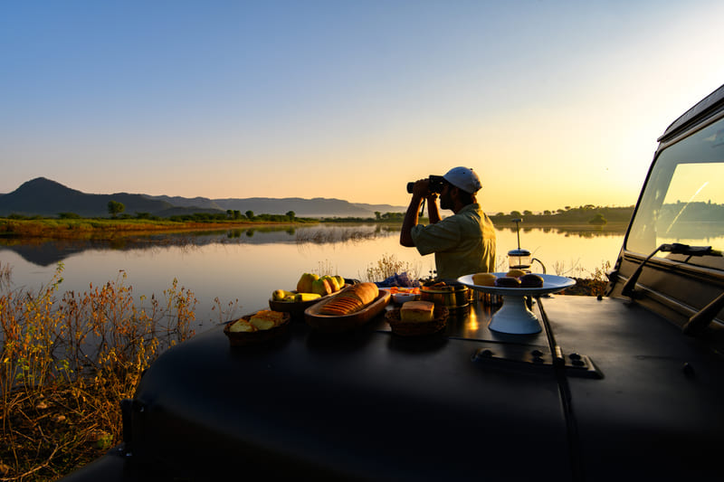 An image of a man standing near a lake with his jeep parked over looking the mountain - An image of a jeep parked amidst the rocky mountains - Utsav Camp Sariska