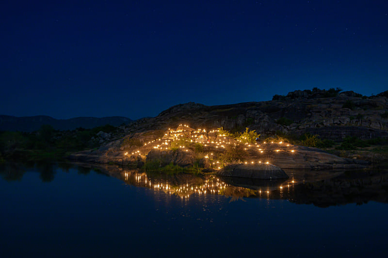 A view of an outdoor dining arrangement near a lake over viewing the mountains with bright lighting and greenery around at Utsav Camp Sariska.