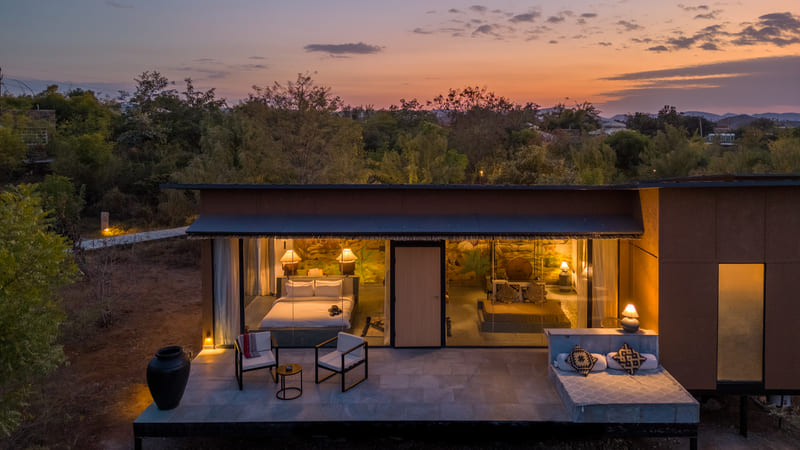 Aerial view of the Forest Bungalow at Utsav Camp Sariska with large glass walls revealing an lit bedroom at dusk.