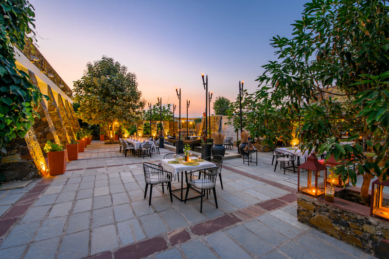 Outdoor dining area at Utsav Camp Sariska with tables and chairs set up on a stone patio surrounded by trees at dusk.