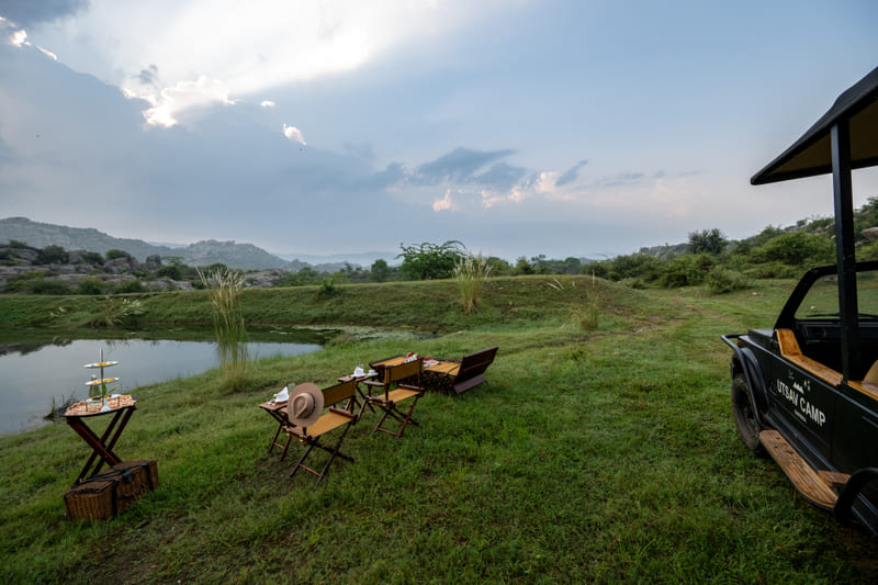Outdoor chairs, a table, and a safari vehicle set up on a grassy lake bank under a cloudy sky at Utsav Camp Sariska.