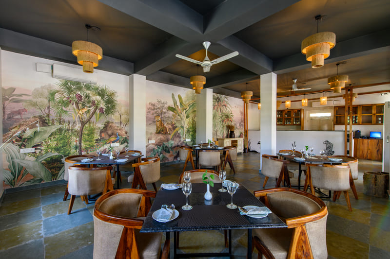 Indoor restaurant at Utsav Camp Sariska featuring wooden tables, chairs, unique hanging lights, and wall murals of nature.