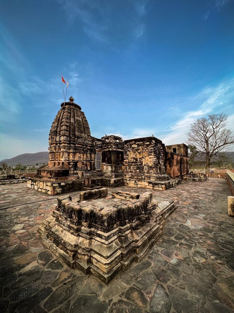 An ancient carved stone temple stands majestically on a rocky platform beneath a bright, cloudless blue sky.