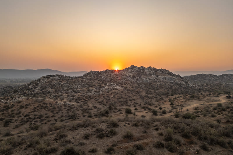 The sun sets behind a rocky hill, casting a warm golden glow across a dry, brush-filled landscape.