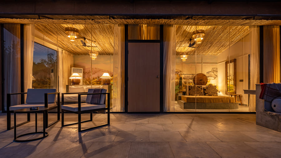 A Forest Bungalow at Utsav Camp Sariska with floor-to-ceiling glass walls showing a lit bedroom and a patio with chairs.