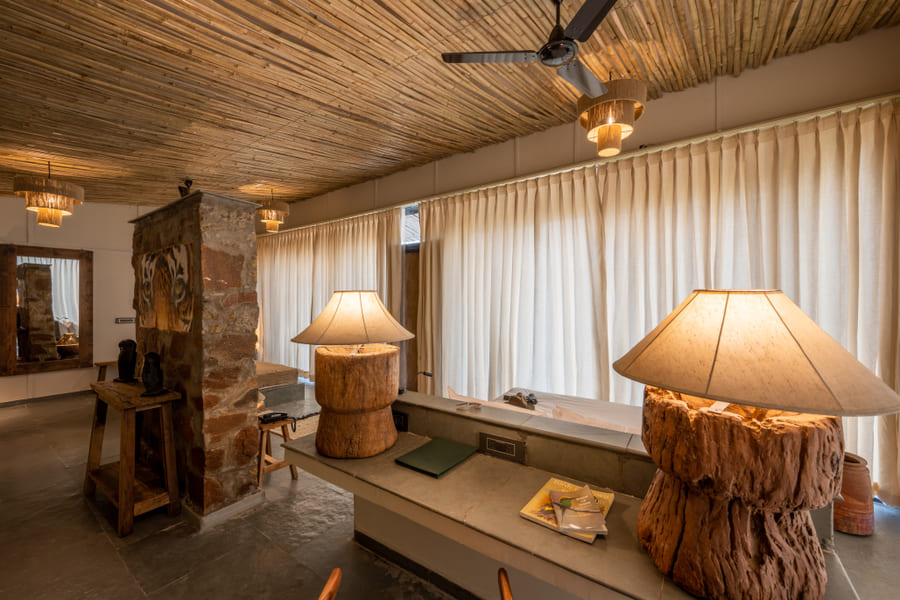 A guest room at Utsav Camp Sariska featuring lamps on unique wooden bases, a desk, a stone wall divider, and long curtains.