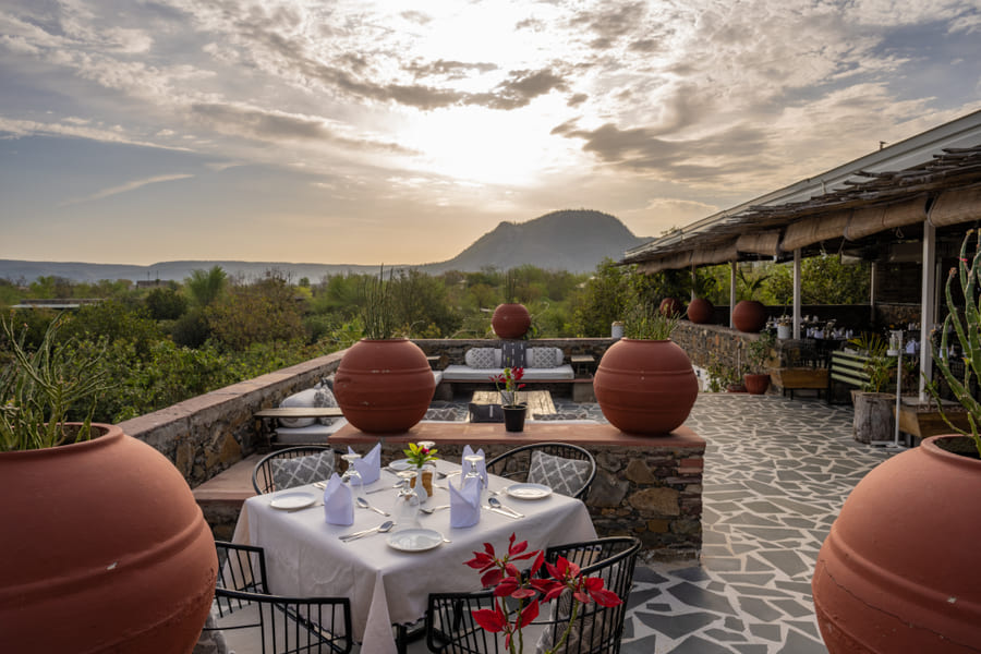 An outdoor rooftop dining terrace at Utsav Camp Sariska featuring set tables and large clay pots, overlooking a valley and hills.