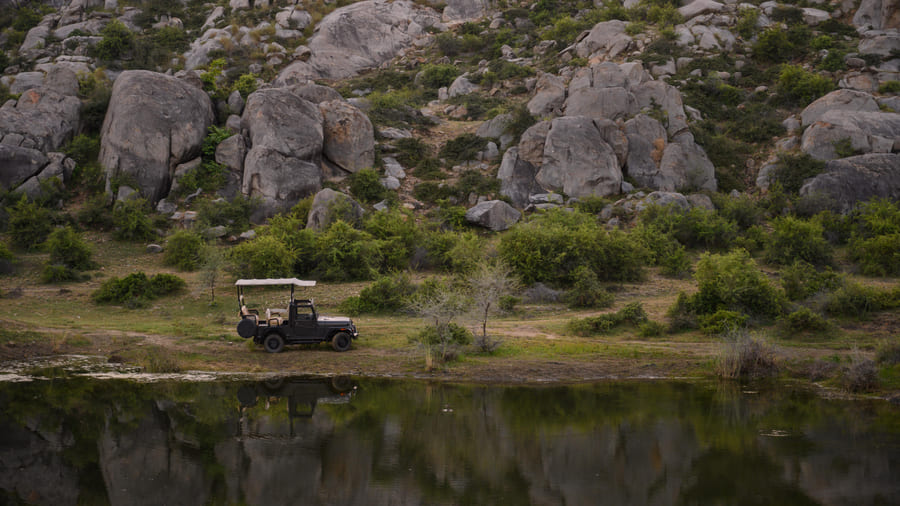 A safari jeep drawn up beside a waterhole below a boulder hillside