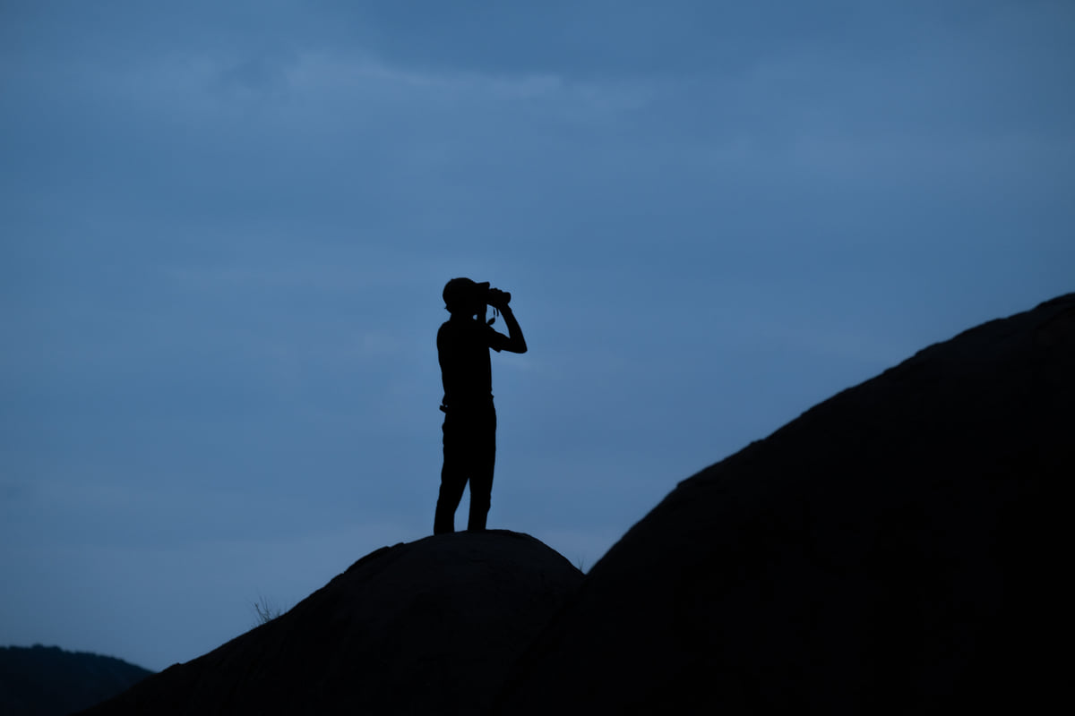 A guest silhouetted on a boulder at dusk, scanning the sky through binoculars