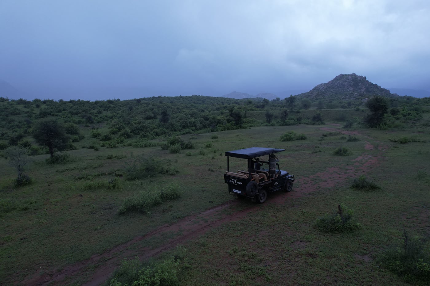 A jeep driving amidst a misty grassland at Utsav Camp Sariska seen with mountains in the background and a cloudy sky at possibly dawn.