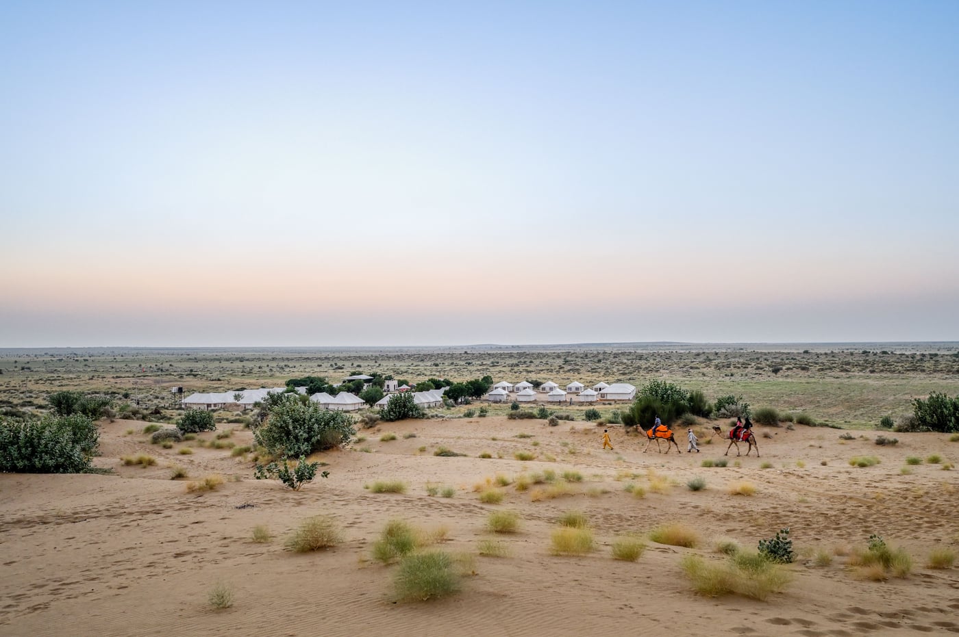 A panoramic view of a vast desert landscape featuring small sand dunes and scattered green bushes at dusk.