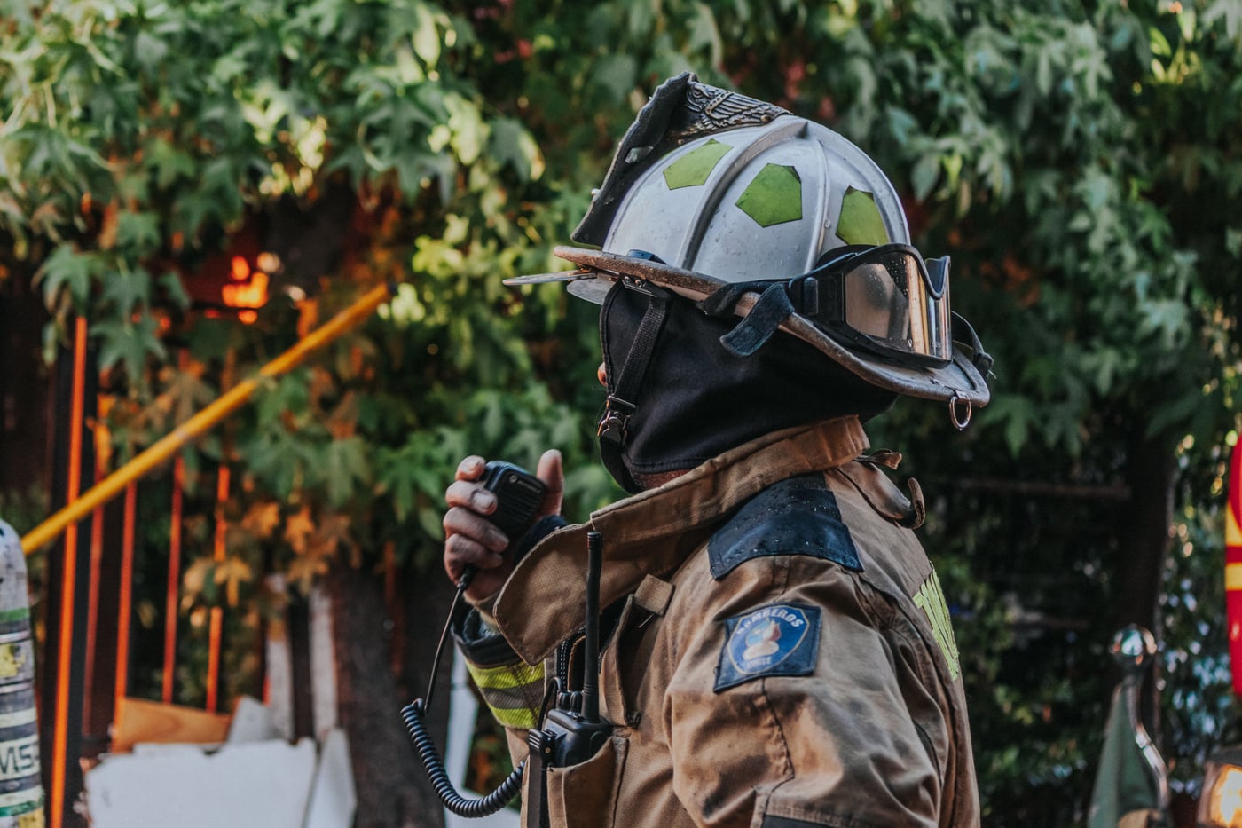 A person wearing a protective helmet and gear holds a walkie-talkie while looking to the side outdoors.