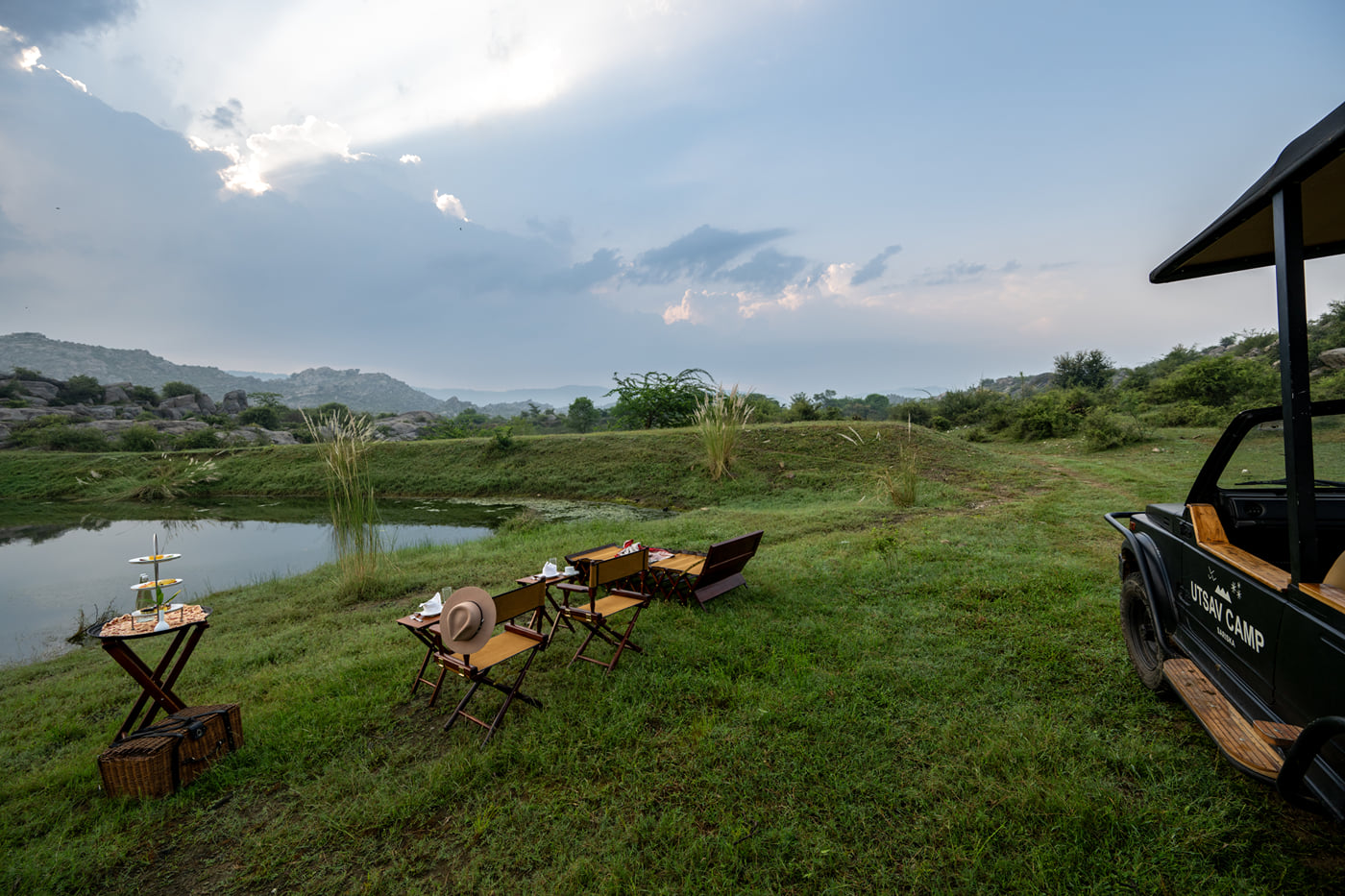 Outdoor chairs, a table, and a safari vehicle set up on a grassy lake bank under a cloudy sky at Utsav Camp Sariska.