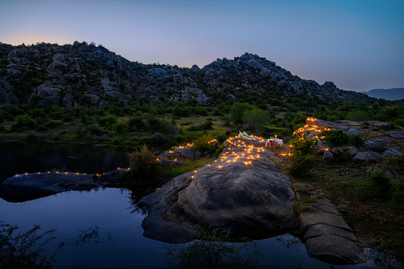 An image of an outdoor dining arrangement near a lake over viewing the mountains with bright lighting and greenery around - Utsav Camp Sariska