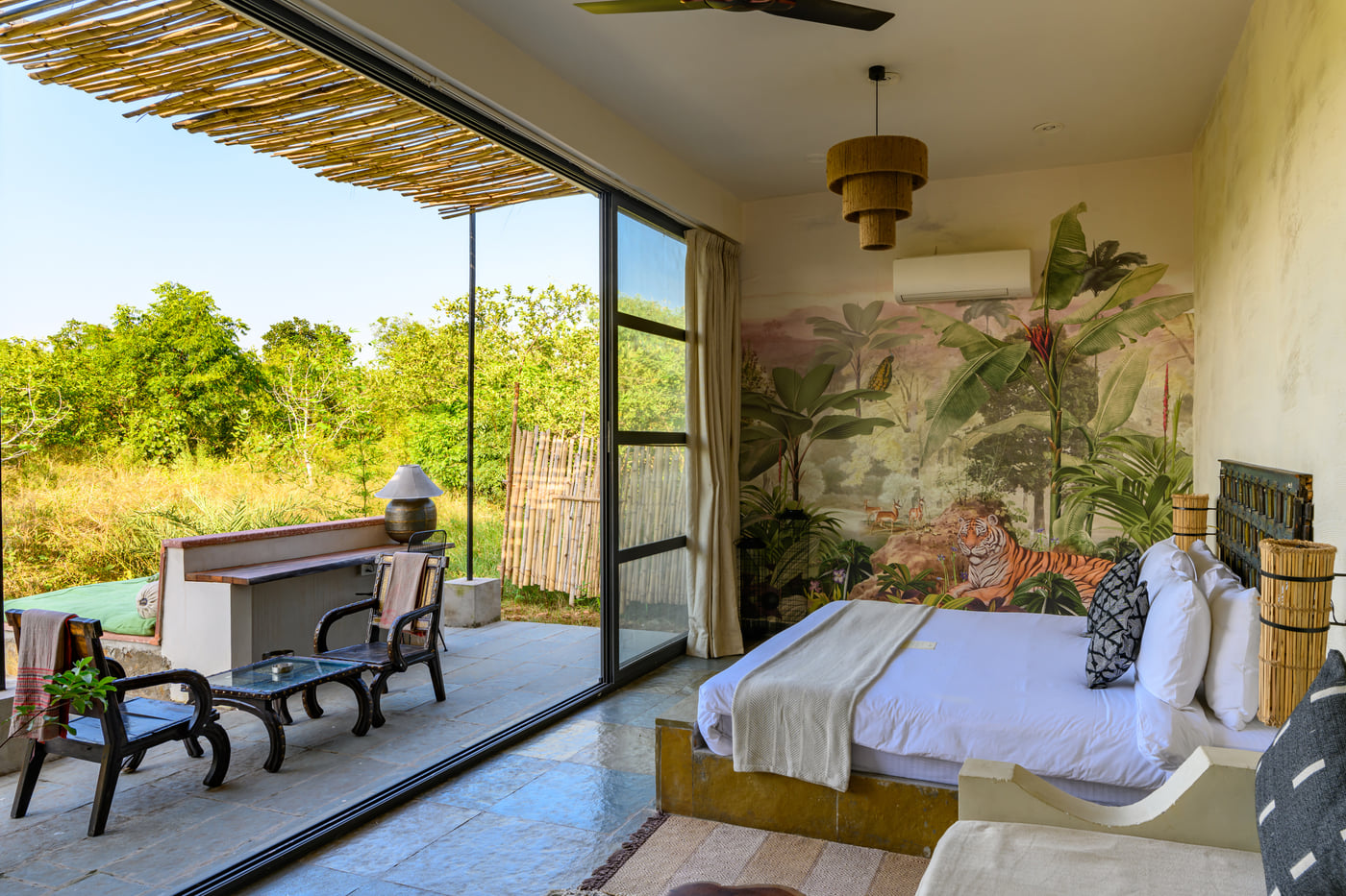 An image of a room with a queen size bed and decorative items with wall hangings and an expansive glass door with an outdoor seating arrangement - Utsav Camp Sariska