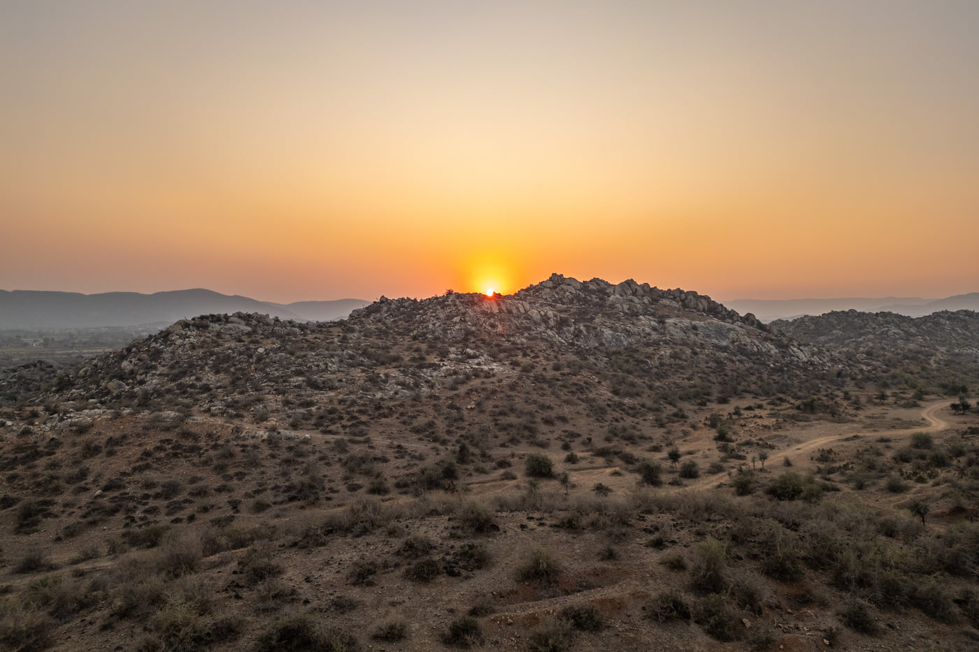 The sun sets behind a rocky hill, casting a warm golden glow across a dry, brush-filled landscape.