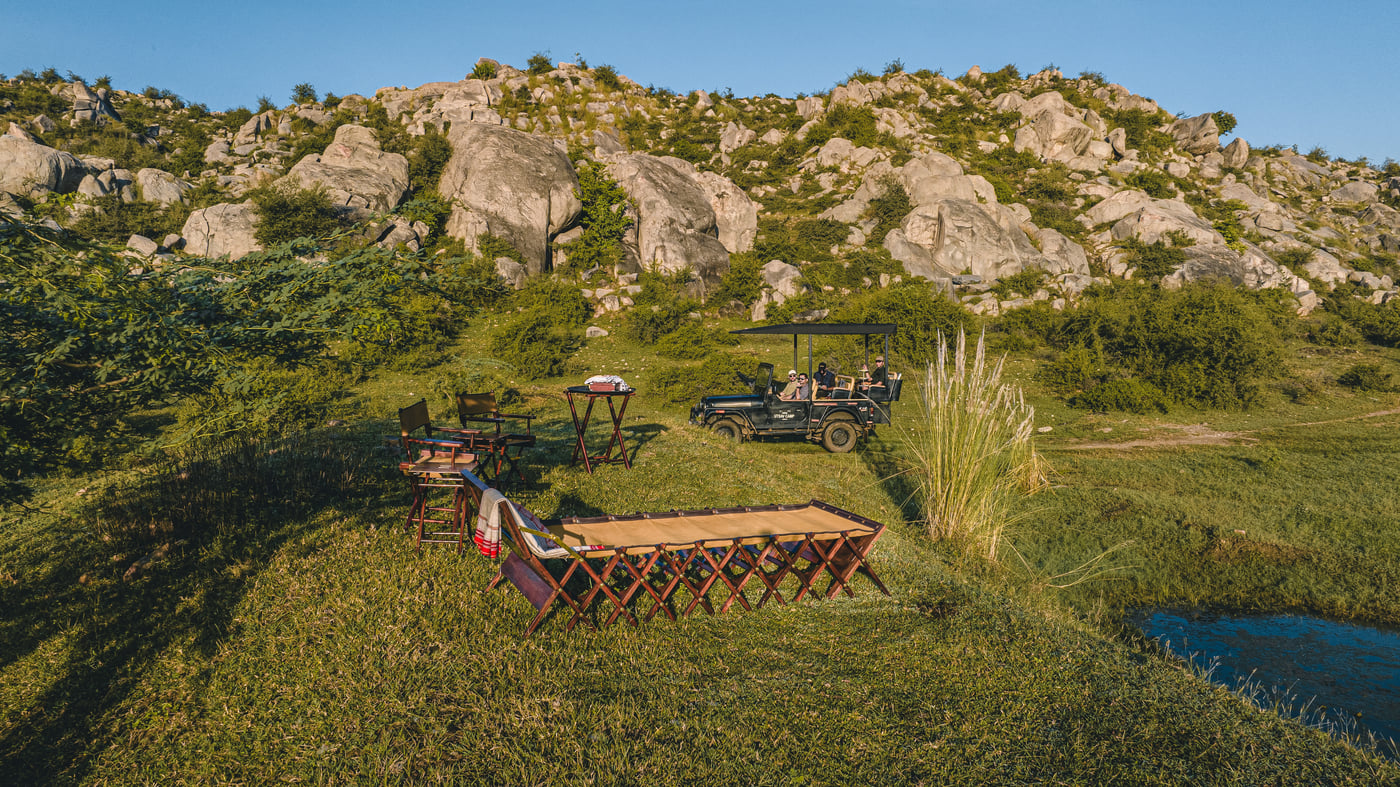 An overview of a sundowner setup at Utsav Camp Sariska by a small waterbody with a lounger, chairs and tables placed on an elevated place and a jeep safari with people inside looking on.