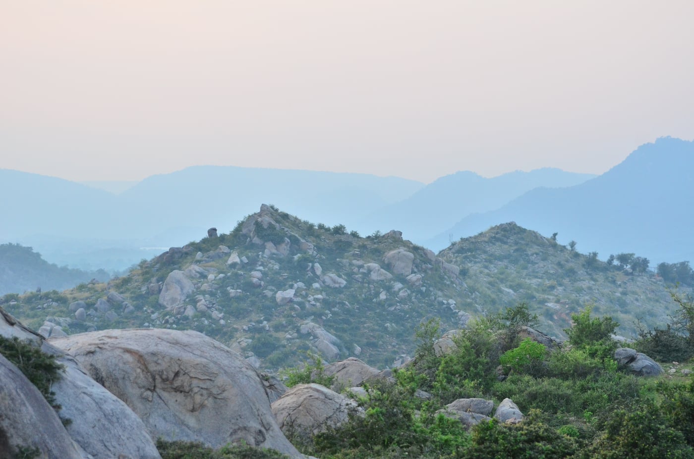 Hazy view of rocky hilltops covered in scattered shrubs and boulders, with layered mountain ridges in the background during dawn or dusk.