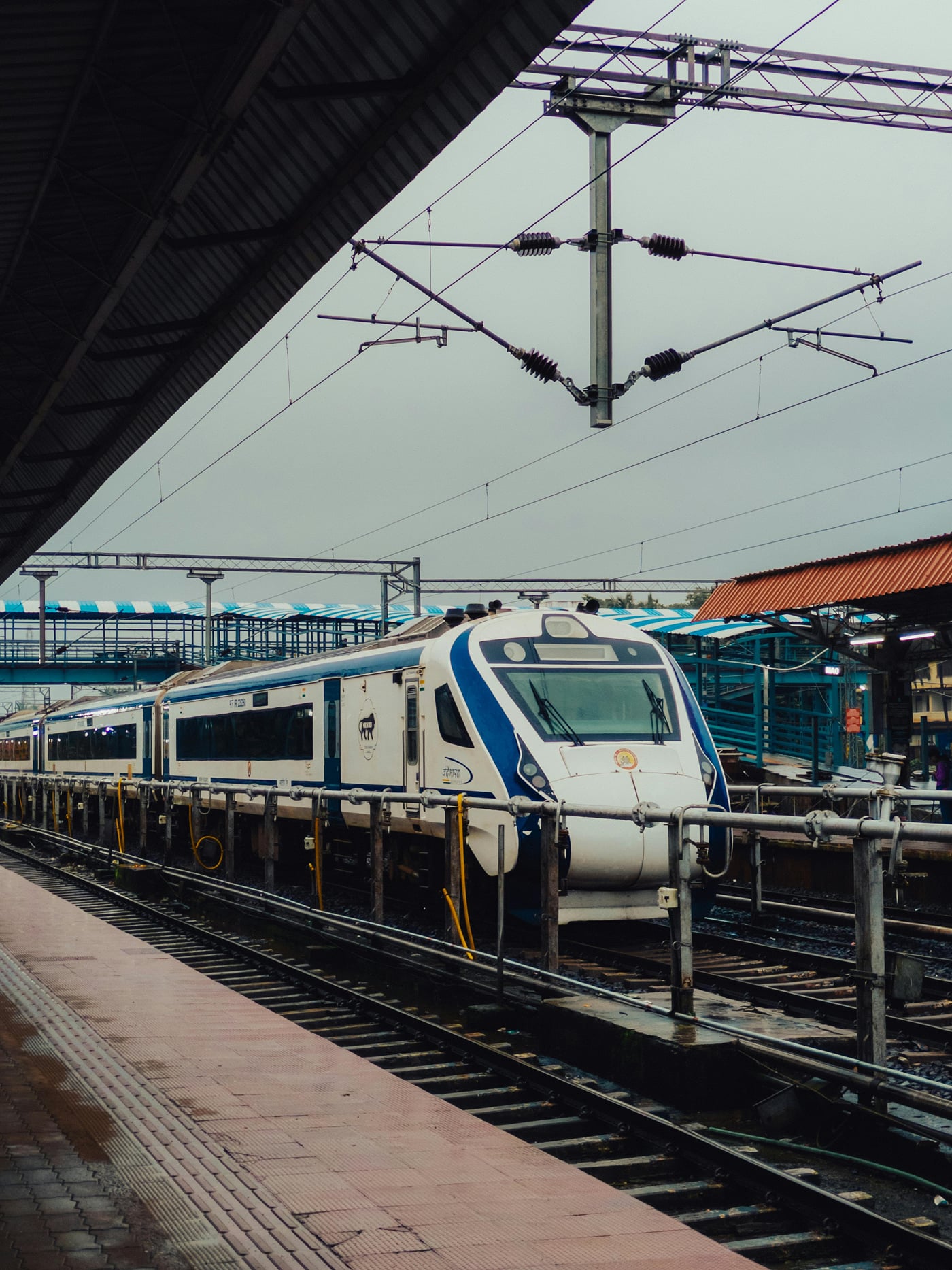 A white and blue Vande Bharat Express train at a railway station platform under overhead electric lines.