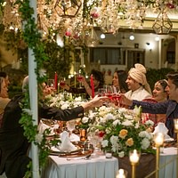 An elegant outdoor dining setup at night, featuring a long table with white tableware and floral arrangements, under cascading fairy lights and surrounded by lush greenery.
