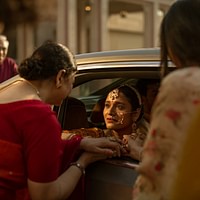 A joyful scene of a woman arriving in a car greeted by two other women and onlookers; they appear engaged in a warm, celebratory interaction, possibly a cultural event.