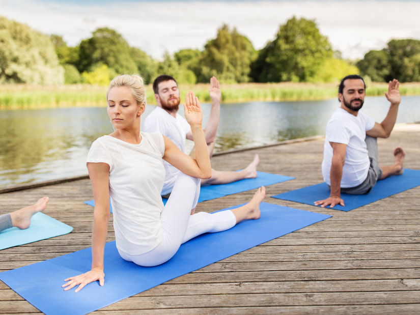 Three people on blue yoga mats on a wooden deck beside a lake at YO1 Longevity & Health Resorts, seated with one arm raised