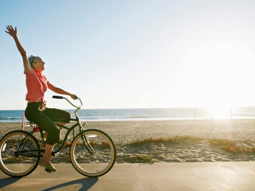 A woman riding a cruiser bicycle along a beach path at sunset with one arm raised