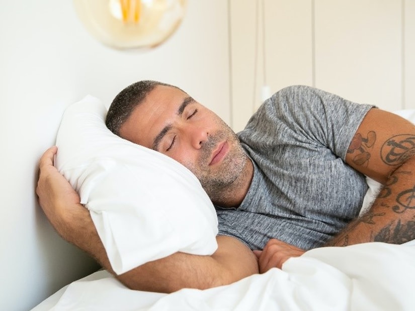 A man asleep on his side, arms wrapped around a white pillow under a warm bedside light