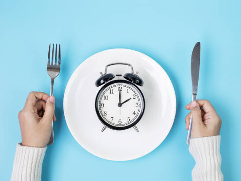 Hands holding a fork and a knife either side of a white plate with a black alarm clock on it, on a pale blue background
