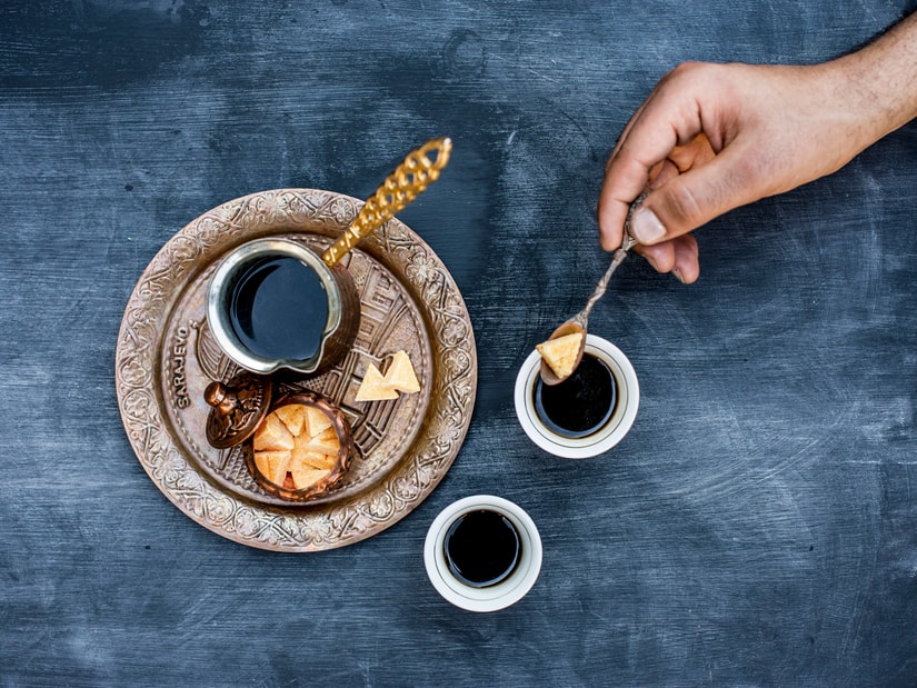 A hand drizzling honey from a spoon into a small cup of black coffee on a brass tray, with a second cup and biscuits on a dark table