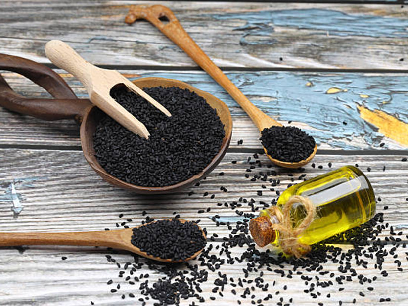 Black kalonji seeds in a wooden bowl and spoons, with a small corked bottle of golden black seed oil on weathered wood