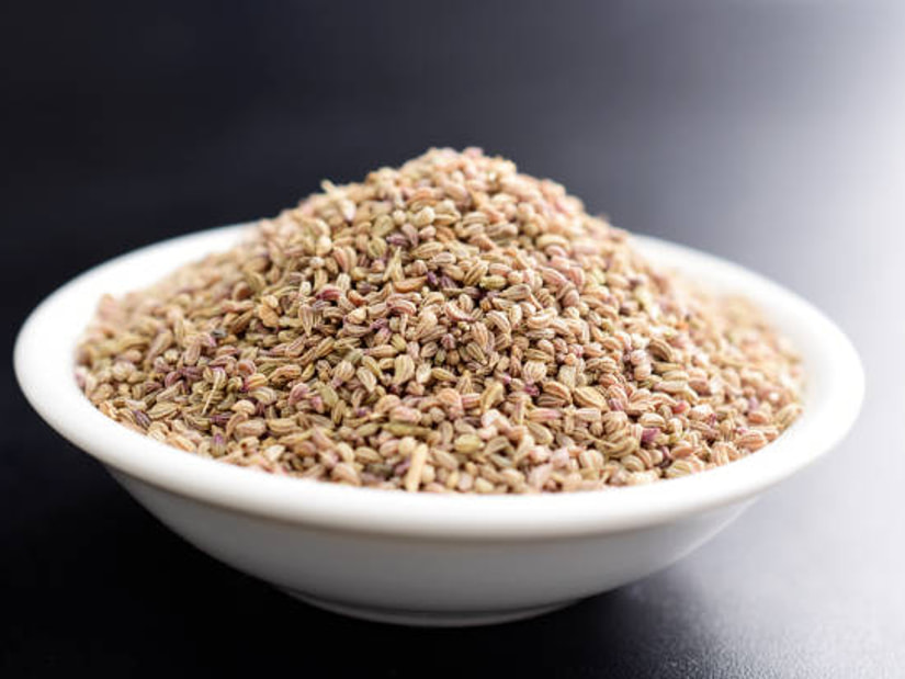A white bowl heaped with ajwain carom seeds on a dark surface