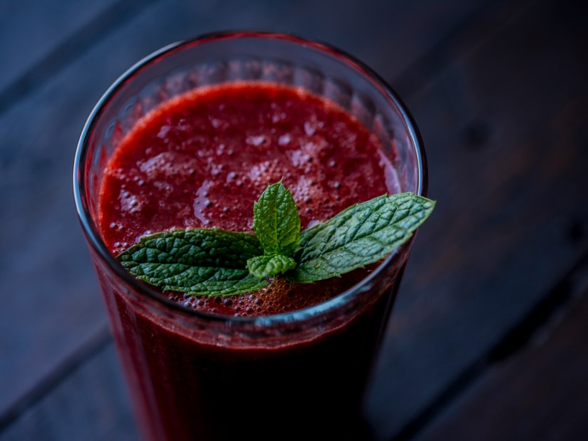 A tall glass of deep red carrot and beetroot juice garnished with a sprig of mint on a dark wooden table, seen from above