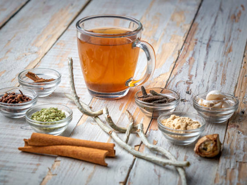 A glass mug of amber herbal decoction surrounded by dried giloy stems, cinnamon sticks, cloves and small bowls of spices
