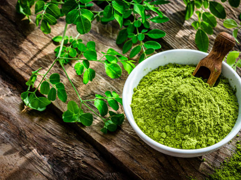 A white bowl of bright green moringa leaf powder with a wooden scoop, beside fresh moringa leaves on weathered wood