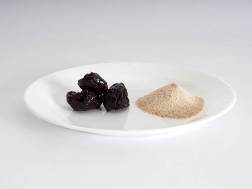 A white plate holding a small mound of pale psyllium husk beside a few dark prunes