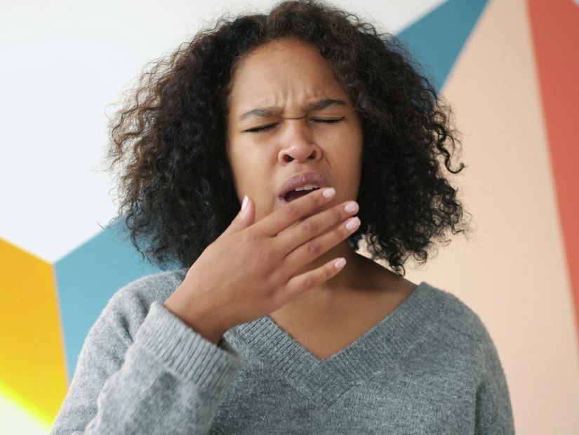 A woman with curly hair yawning with her hand to her mouth against a brightly painted geometric wall