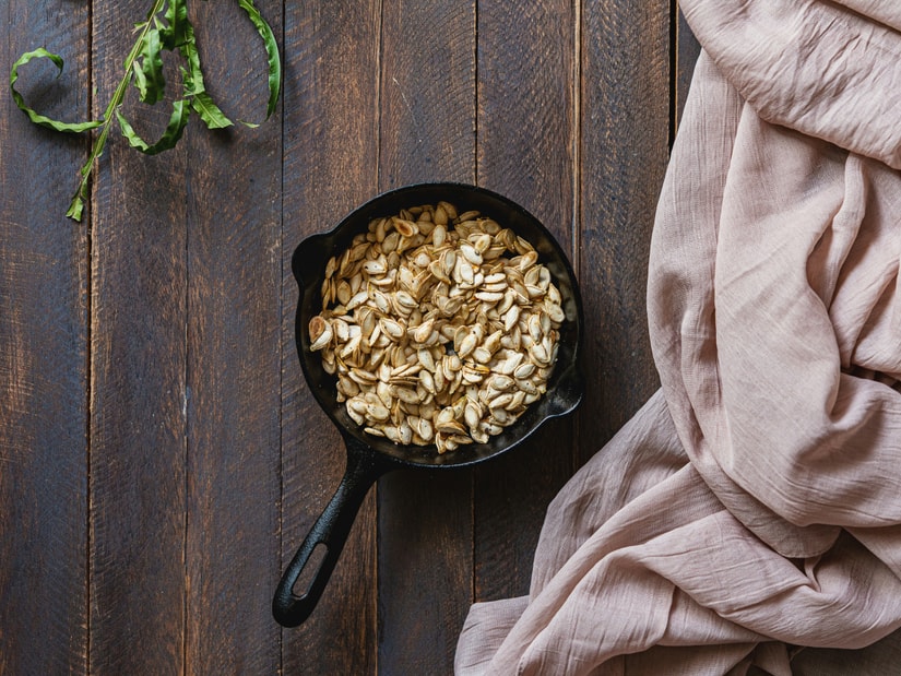 A small cast iron skillet filled with roasted pumpkin seeds on a dark wood surface with a dried leaf and blush-pink linen nearby