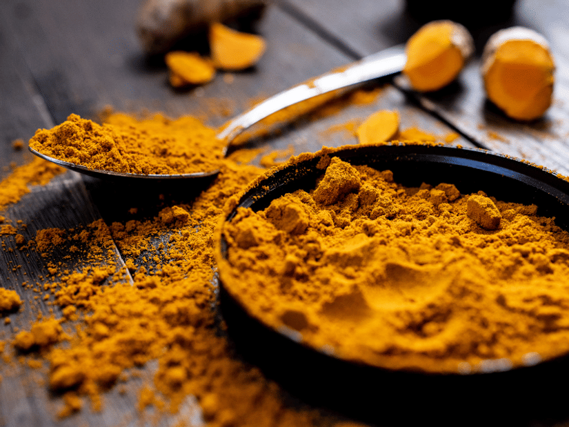 A spoon and small dish overflowing with ground turmeric powder next to fresh turmeric root pieces on a dark wooden surface