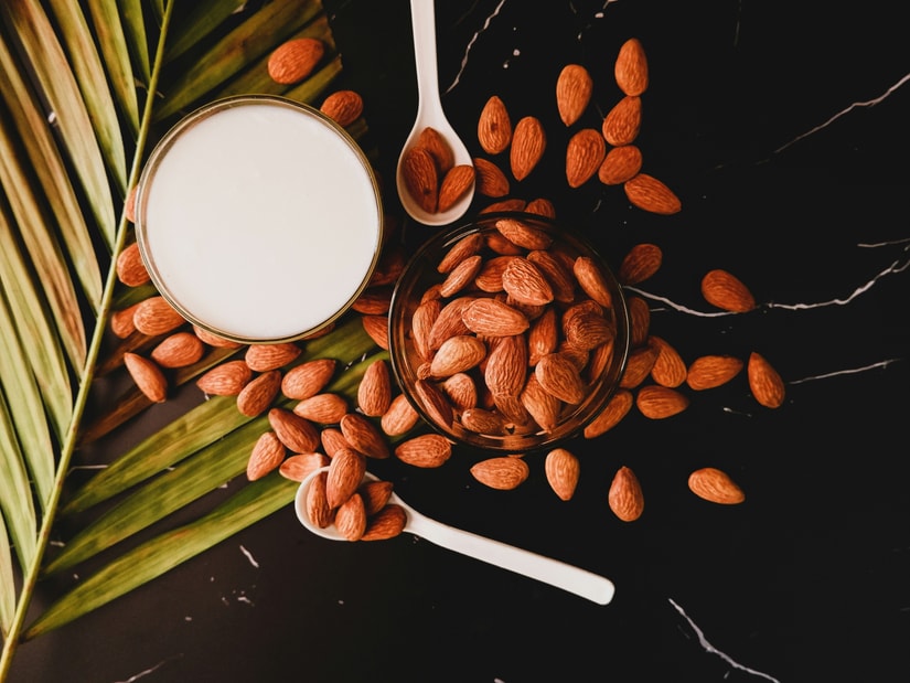 Almonds scattered on a dark marble surface with a bowl of almonds, a glass of almond milk, two white spoons and a palm leaf, seen from above