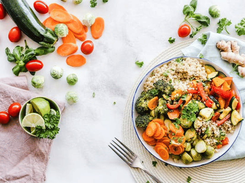 An overhead spread of a quinoa and roasted vegetable bowl surrounded by raw vegetables, ginger and lime