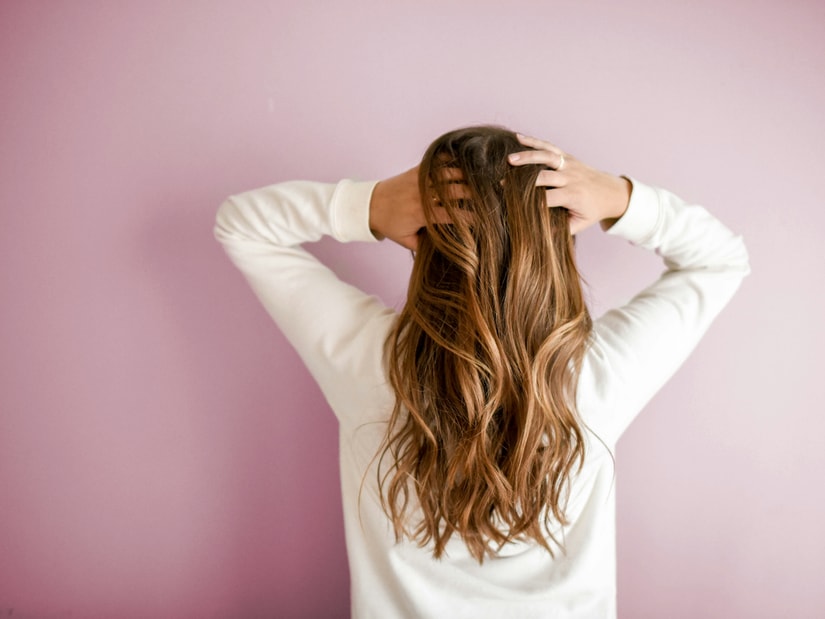 A woman seen from behind against a pink background, running both hands through long wavy hair