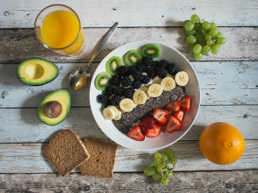 A bowl of fruit and chia seeds beside avocado halves, grapes, an orange, rye bread and a glass of juice on a wooden table