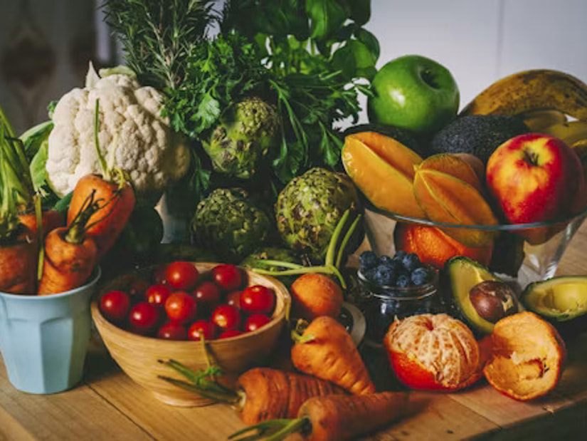 A wooden table laid with cauliflower, artichokes, carrots, cherry tomatoes, avocado and fresh fruit