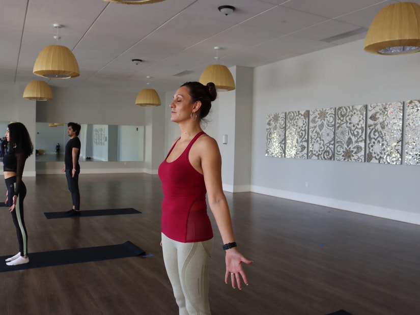 A woman standing with her eyes closed and palms open in a yoga studio, two people on mats behind her