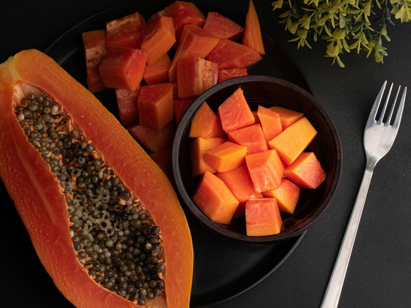 A halved papaya showing its cluster of black seeds, beside a plate and a wooden bowl of cubed papaya and a fork on a dark surface