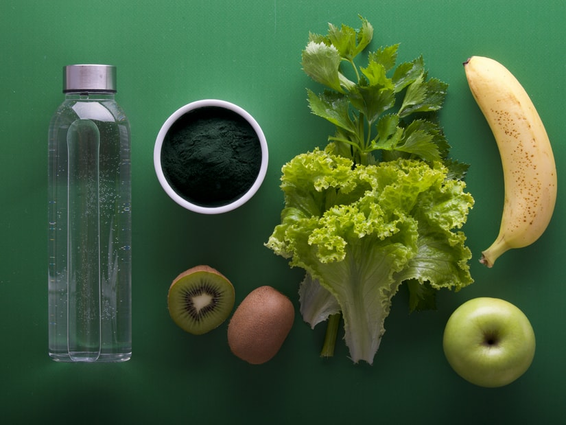 A flat lay on a green background of a glass water bottle, a bowl of green powder, leafy lettuce, a banana, halved kiwi, an egg and a green apple